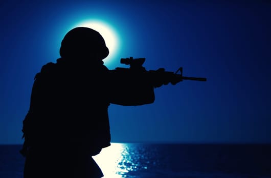 Silhouette of army special forces rifleman, soldier in ammunition and helmet aiming gun, shooting with service rifle on seacoast at night. Commando fighter, marine raider during amphibious mission