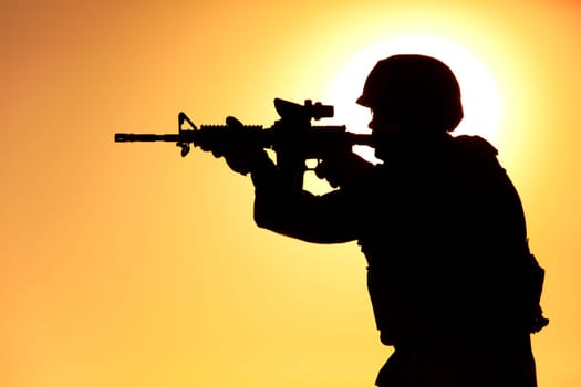 Silhouette of modern army infantry soldier standing on background of setting sun and aiming assault rifle with collimator sight. Special operations rifleman, elite forces shooter shooting in enemy