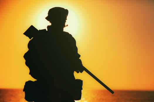 Silhouette of army elite forces sniper, commando shooter in boonie hat, carrying backpack, equipped tactical radio, walking on background of setting or raising sun. Counter terrorist forces soldier