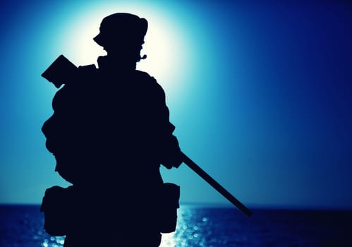 Silhouette of army elite forces fighter standing with sniper rifle on background of blue sky with moon or sun. Sniper or marksman in boonie hat, carrying backpack on mission, patrolling at night
