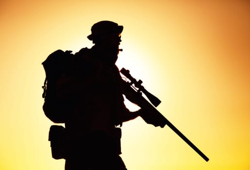 Silhouette of army elite forces sniper, commando shooter in boonie hat, carrying backpack, equipped tactical radio, walking on background of setting or raising sun. Counter terrorist forces soldier