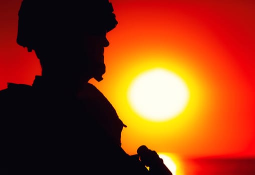 Shoulder silhouette of army soldier, special operations forces fighter in combat helmet and ballistic glasses, standing with gun on background of setting below ocean horizon sun