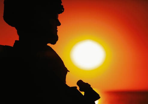 Shoulder silhouette of army soldier, special operations forces fighter in combat helmet and ballistic glasses, standing with gun on background of setting below ocean horizon sun