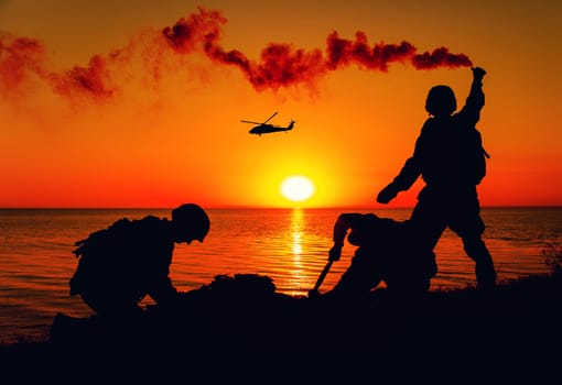 Silhouette of army special forces infantry soldiers, Marines or Navy SEALS team signaling to helicopter with smoke flair while waiting for evacuation, landing on seashore during amphibious operation