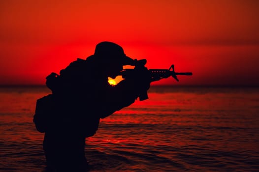 Silhouette of army soldier aiming and shooting service rifle while standing knee-deep in sea water. Coast guard, naval forces soldier attacking enemy with weapon, landing on beach on sunset or dawn