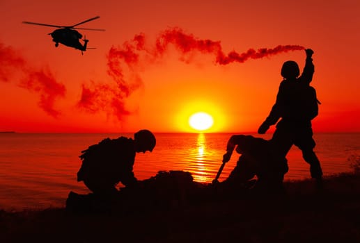 Silhouette of army special forces infantry soldiers, Marines or Navy SEALS team signaling to helicopter with smoke flair while waiting for evacuation, landing on seashore during amphibious operation