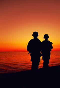 Silhouette of two Marines soldiers, special operations infantrymen or coast guard fighters in uniform and combat helmet, armed service rifles, standing on seashore, patrolling beach at sunset or dawn