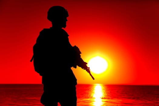 Silhouette of army special forces rifleman, Marines, coast guard or anti-terrorist squad soldier in ammunition and helmet, armed service rifle, standing on ocean shore, patrolling seacoast at sunset