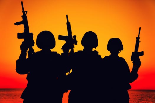 Silhouette of army special operations forces soldiers team, group of Marines or coast guard fighters crew in ammunition and combat helmets standing on seashore at sunset with raised assault rifles