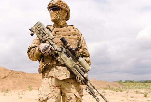 Portrait of armed special forces soldier with sniper rifle standing against dramatic sky
