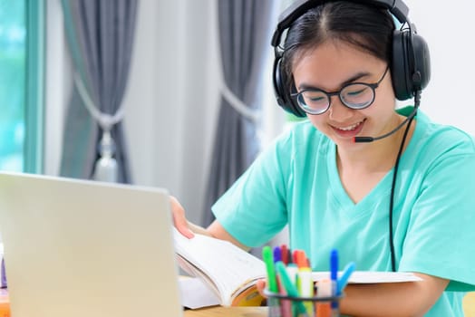 Portrait young Asian woman student with headphones glasses is smiling reading a book study a teenage girl is happy to college internet distance learning online class works on a laptop computer at home