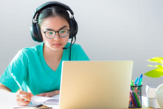 Asian young woman student with glasses headphones study serious writing note on a book looking video conference on laptop computer university class online internet learning distance education at home