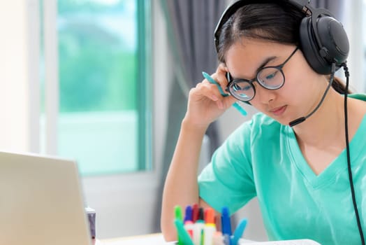 Asian woman student teenage girl with glasses headphones sitting looking serious reading a book worry using laptop computer on table learning online study. Education from a class of university at home