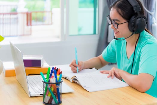 Asian young woman student with glasses headphones girl study happy writing note on a book looking video conference laptop computer university class online internet learning distance education at home