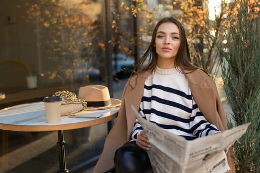 Beautiful girl in a coat reads a newspaper and smiles while relaxing in an outdoor cafe.