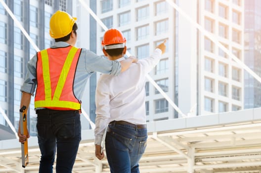 Back view of two male wearing safety helmet and looking at modern building. Construction foreman wearing orange safety vest with yellow reflective stripe and holding spirit level while project engineering manager holding laptop and pointing finger to construction building for successful project. Construction and engineering concept.