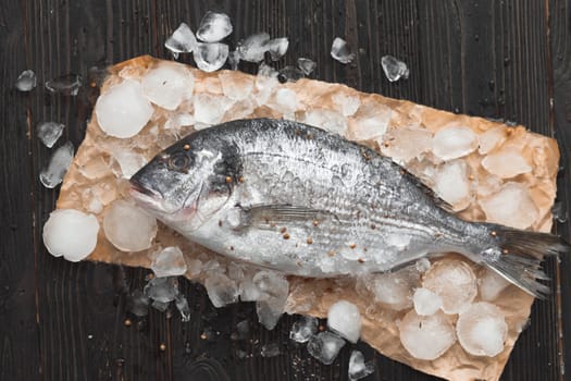 Raw dorada fish or gilt-head bream on ice with lemon slices and rosemary over black wooden background, flat lay, top view.