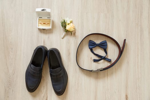 Groom set of accessories flat layout on wooden floor. Top view of bow-tie, belt, shoes, floral corsage, golden rings in wooden box