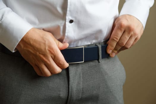 A man in blue pants and a white shirt buttoned a brown leather trouser belt. He has a watch on his hand.