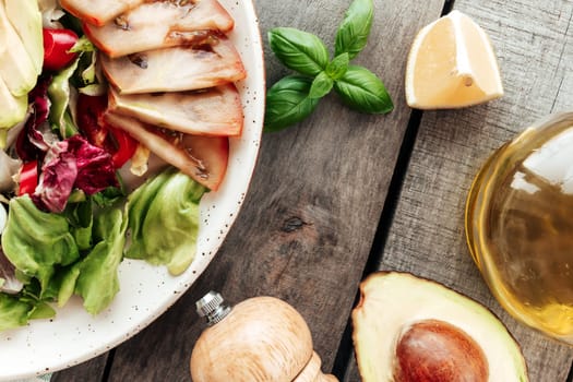 Healthy eating concept flat lay. Mediterranean diet, plate with lettuce salad leaves, avocado half with kernel, pepper, black tomato slices, basil, olive oil, lemon, salt on wooden background