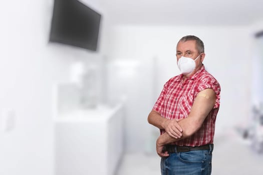 Coronavirus protective measures. A senior man in medical mask shows plasters on his upper arm after Covid-19 vaccination against the blurred background of a doctor's office.