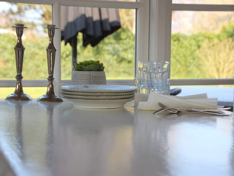 A vintage white table about to be set, with plates, glasses, and cutlery. A lawn can be seen in the background.