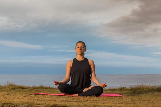 Middle aged woman in black tank top sitting on a mat doing meditation outdoors with sea and cloudy sky in background. Caucasian woman relaxing in yoga pose.