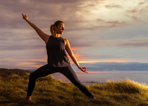 Middle-aged woman in black sportswear doing meditation in front of the sea at sunset. Caucasian woman in yoga pose at dusk.