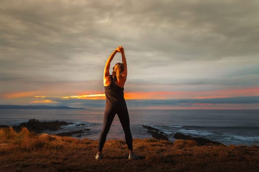 Middle-aged woman in black sportswear on her back stretching in front of the sea at sunset. Caucasian woman relaxing at dusk.
