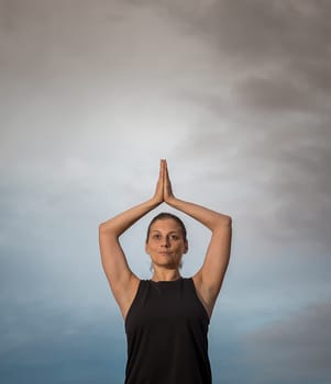 Middle-aged woman in black shirt with her arms over her head doing meditation outdoors with cloudy sky in the background. Caucasian woman relaxing in yoga pose