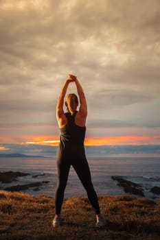 Middle-aged woman in black sportswear on her back stretching in front of the sea at sunset. Caucasian woman relaxing at dusk.