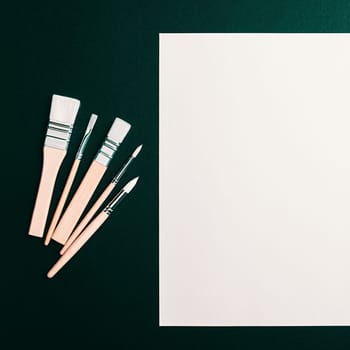 A clean white sheet and paint brushes on a dark green background with space to copy. Mock-up, mockup, layout.