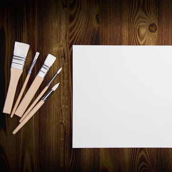 A clean white sheet and brushes on a wood textured background with space to copy. Layout, mockup space for text and drawing.