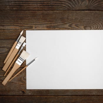 A clean white sheet and brushes on a wood textured background with space to copy. Layout, mockup space for text and drawing.