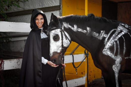 A beautiful girl in medieval style clothes looks happily into the frame and feeds a horse painted with white paint with a painted skeleton
