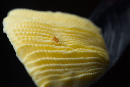 Serrated knife holds a fan shaped curl of rippled yellow margarine. Isolated on black background and a super macro perspective. Center bread crumb too