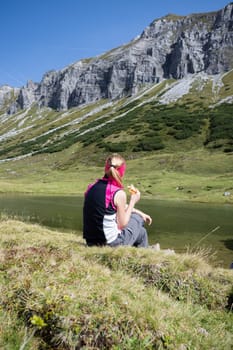 Woman in sportswear is doing a break during a hike: sitting on the ground and enjoying the view over the mountains and a lake