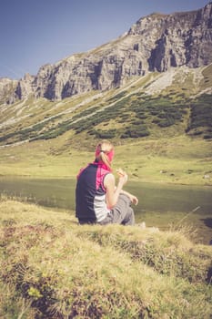 Woman in sportswear is doing a break during a hike: sitting on the ground and enjoying the view over the mountains and a lake
