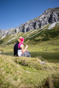 Woman in sportswear is doing a break during a hike: sitting on the ground and enjoying the view over the mountains and a lake