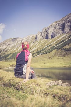Woman in sportswear is doing a break during a hike: sitting on the ground and enjoying the view over the mountains and a lake