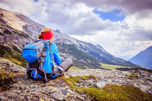 Woman in sportswear and with backpack is sitting on the stony ground and enjoys the view, in the mountains