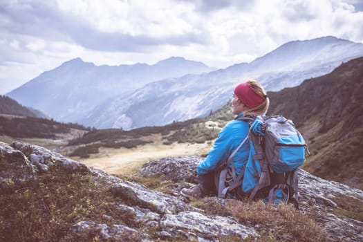 Woman in sportswear and with backpack is sitting on the stony ground and enjoys the view, in the mountains