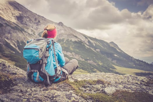 Woman in sportswear and with backpack is sitting on the stony ground and enjoys the view, in the mountains