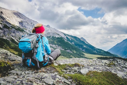 Woman in sportswear and with backpack is sitting on the stony ground and enjoys the view, in the mountains