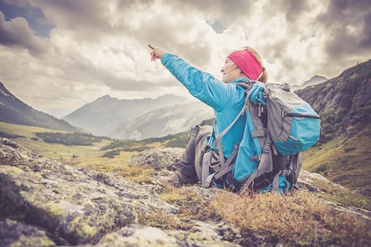 Mountain climber with backpack is sitting on the stony ground and doing a pointing gesture