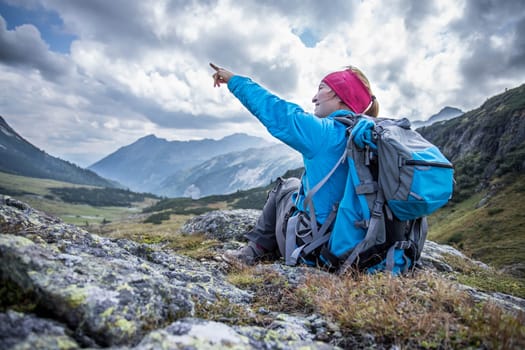 Mountain climber with backpack is sitting on the stony ground and doing a pointing gesture