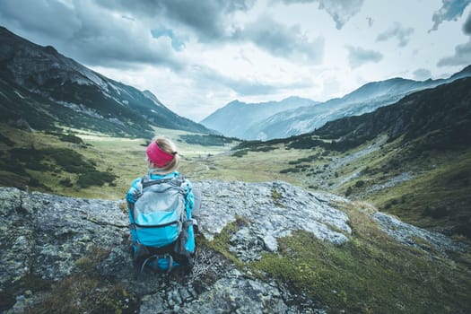 Woman in sportswear and with backpack is sitting on the stony ground and enjoys the view, in the mountains