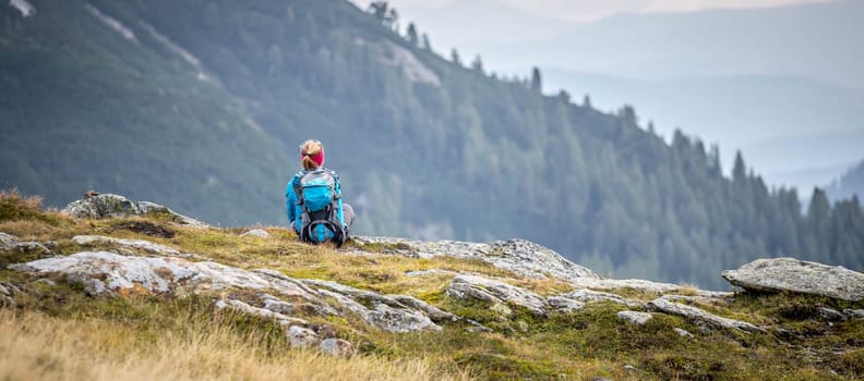 Woman in sportswear and with backpack is sitting on the stony ground and enjoys the view, in the mountains