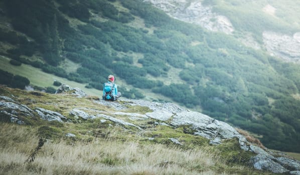 Woman in sportswear and with backpack is sitting on the stony ground and enjoys the view, in the mountains