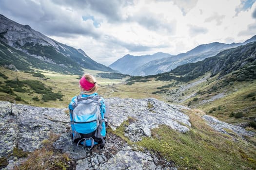 Woman in sportswear and with backpack is sitting on the stony ground and enjoys the view, in the mountains